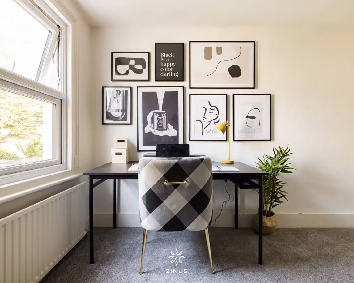 Bright home office with a black desk, checkered upholstered chair, laptop, gold lamp, and a gallery wall of abstract black-and-white art, creating a clean, modern workspace beside a large window and potted plant.