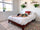 Person in green workout set kneeling in child's pose on a white bed in a bright bedroom with hanging plants and a red pillow.