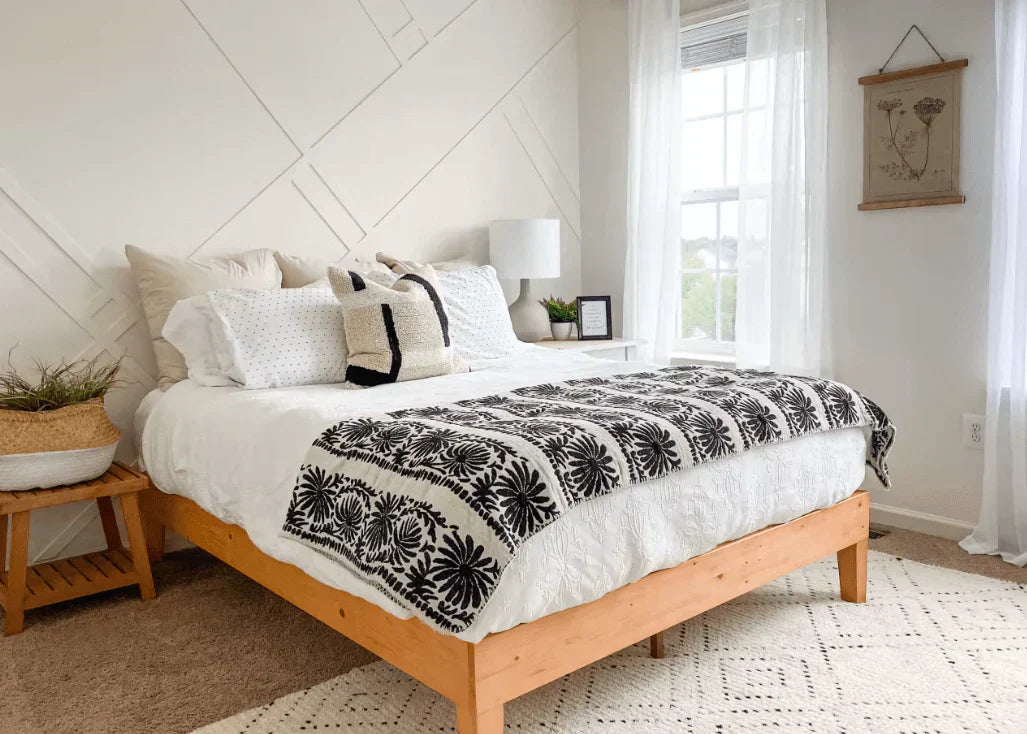 Cozy bedroom with a light wood platform bed, white bedding, textured pillows, and a black-and-white patterned throw, styled with soft natural light and minimal decor.