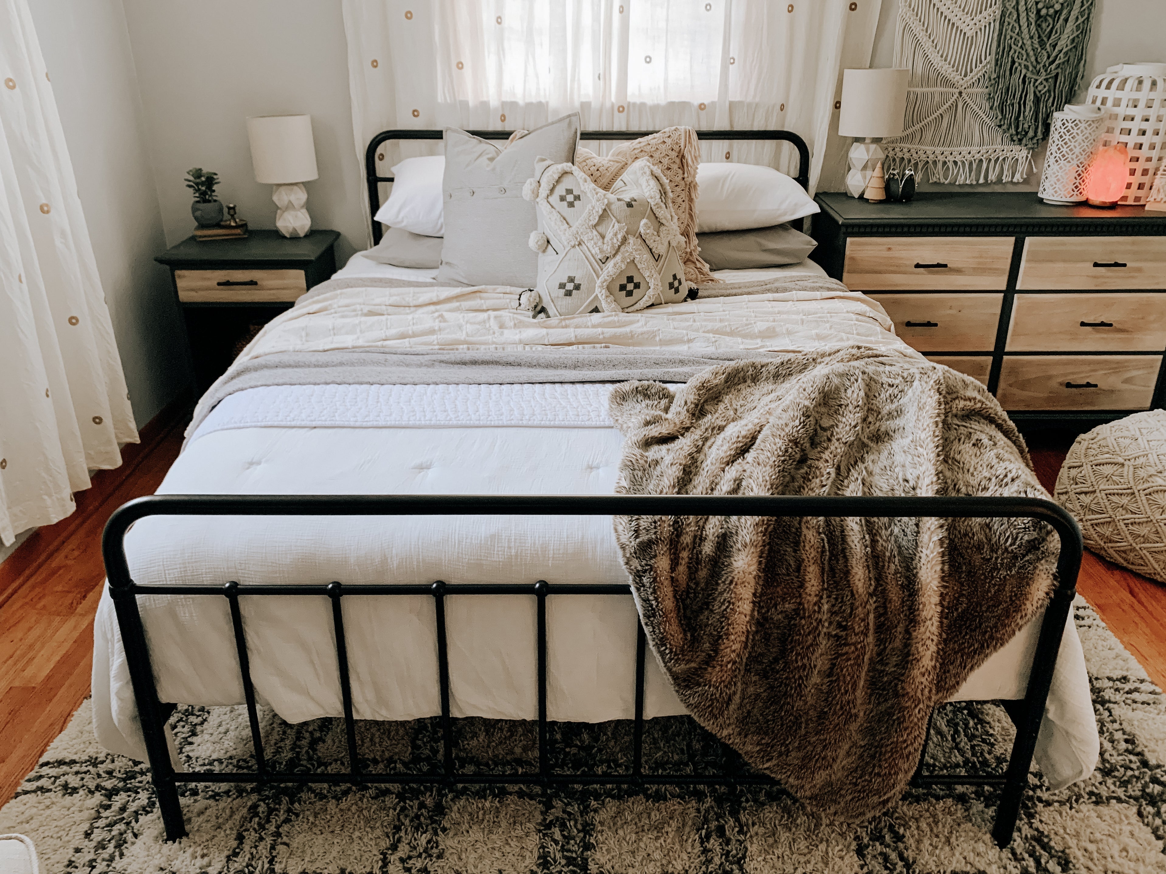 Modern rustic bedroom with black metal bed frame, white bedding, tan blanket, and neutral pillows; wood nightstands, woven decor, and soft natural light create a cozy retreat