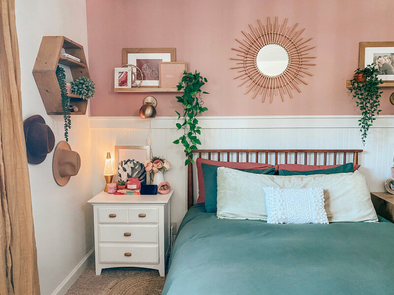 Neutral modern bedroom in beige and white tones featuring a bed with layered patterned pillows, taupe blanket, and a tray with candle and plant; paneled wall, wreath, and white nightstands with flowers and books create a calm, elegant atmosphere.
