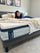Person smiling while lying on a bed showcasing an Ultra Cooling Green Tea mattress with a textured white top and blue cooling label, highlighting comfort and the mattress’s cooling support in a bright bedroom