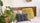Cozy bed with a wooden headboard, decorated with a yellow shag pillow, a black patterned pillow with tassels, and a beige pillow featuring a Buddha illustration, next to large green houseplants and woven baskets for a warm, earthy look.