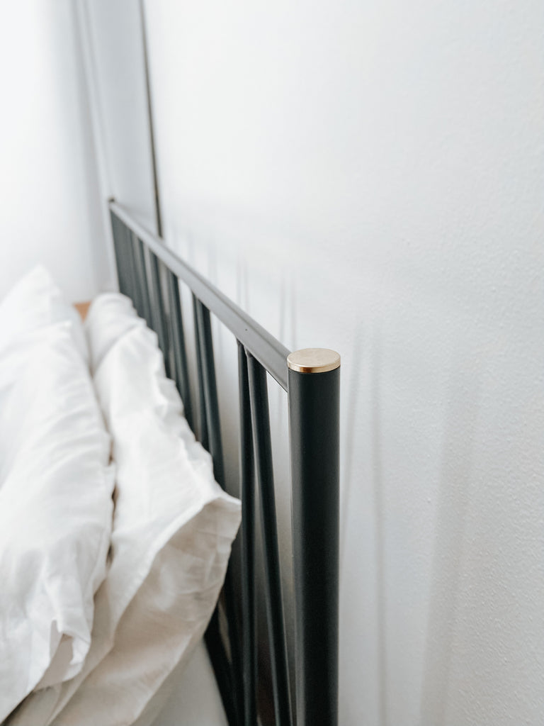 Close-up of black metal bed frame headboard with slim vertical spindles and a rounded wood-accent cap, shown beside white pillows against a light wall to emphasize sturdy construction, modern design, and smooth finish details