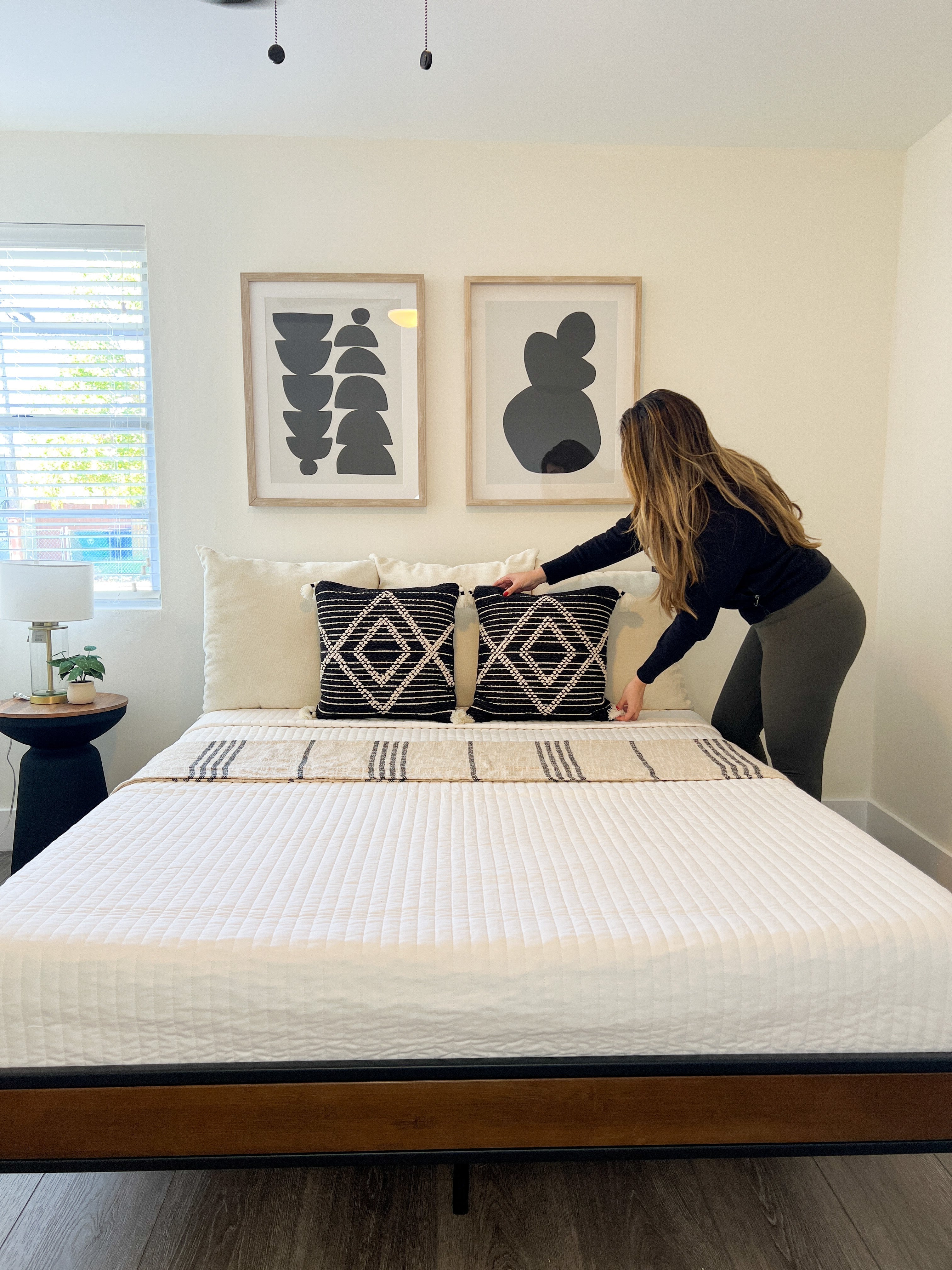 Modern bedroom with a neatly made bed on a Zinus wood-and-metal frame as a person adjusts patterned accent pillows beneath two abstract black-and-white art prints, emphasizing simple styling and clean lines.