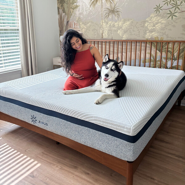 Zinus queen sized cooling memory foam mattress on a wooden platform bed, shown supporting a relaxed person sitting beside a large dog in a bright bedroom to highlight comfort and durability.