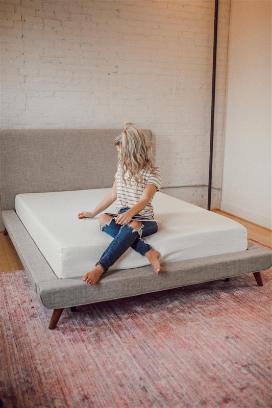 Zinus memory foam mattress on a low gray platform bed in a bright room, shown supporting a seated person to highlight simple comfort and supportive cushioning