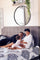 Family relaxing together on a gray upholstered headboard bed with patterned bedding, showing a joyful morning moment and comfortable bedroom setting with a round mirror and greenery.