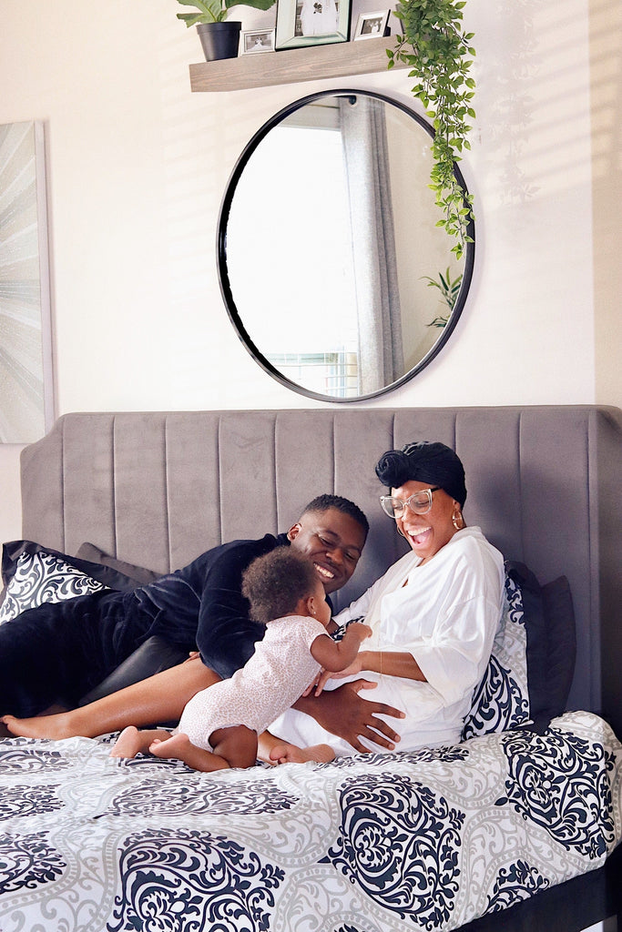 Family relaxing together on a gray upholstered headboard bed with patterned bedding, showing a joyful morning moment and comfortable bedroom setting with a round mirror and greenery.