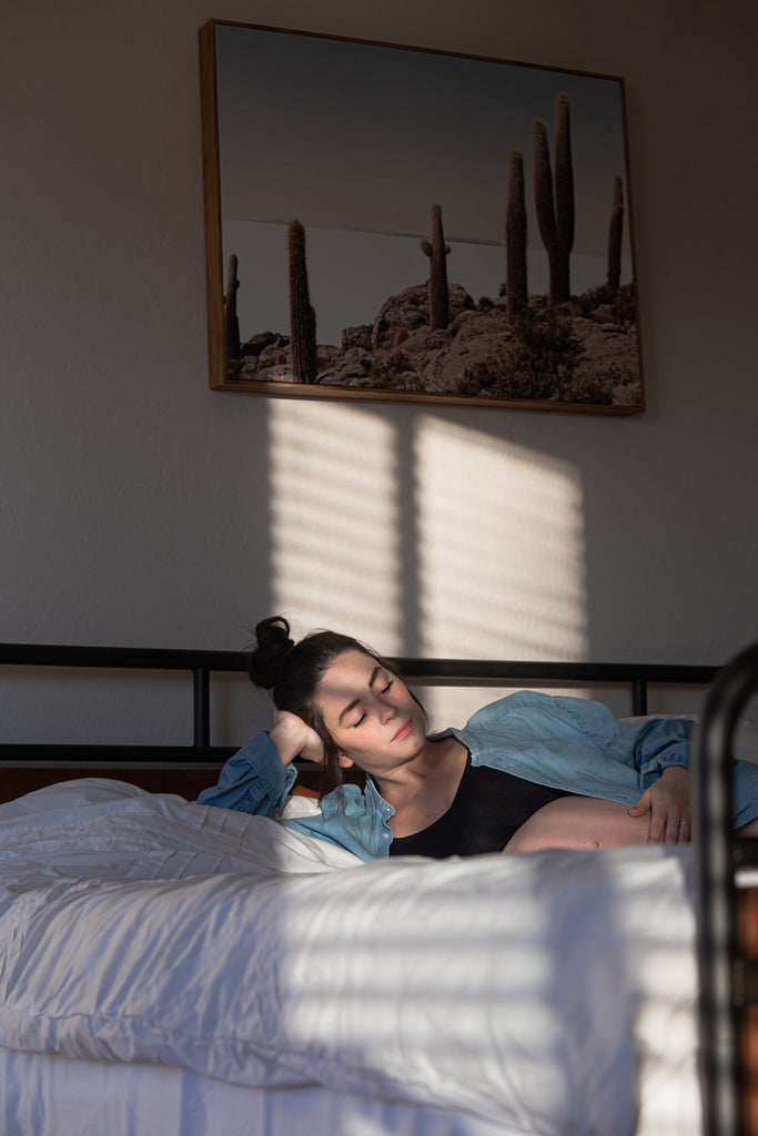 Person resting on a sunlit bed, leaning on one arm beneath cactus artwork.