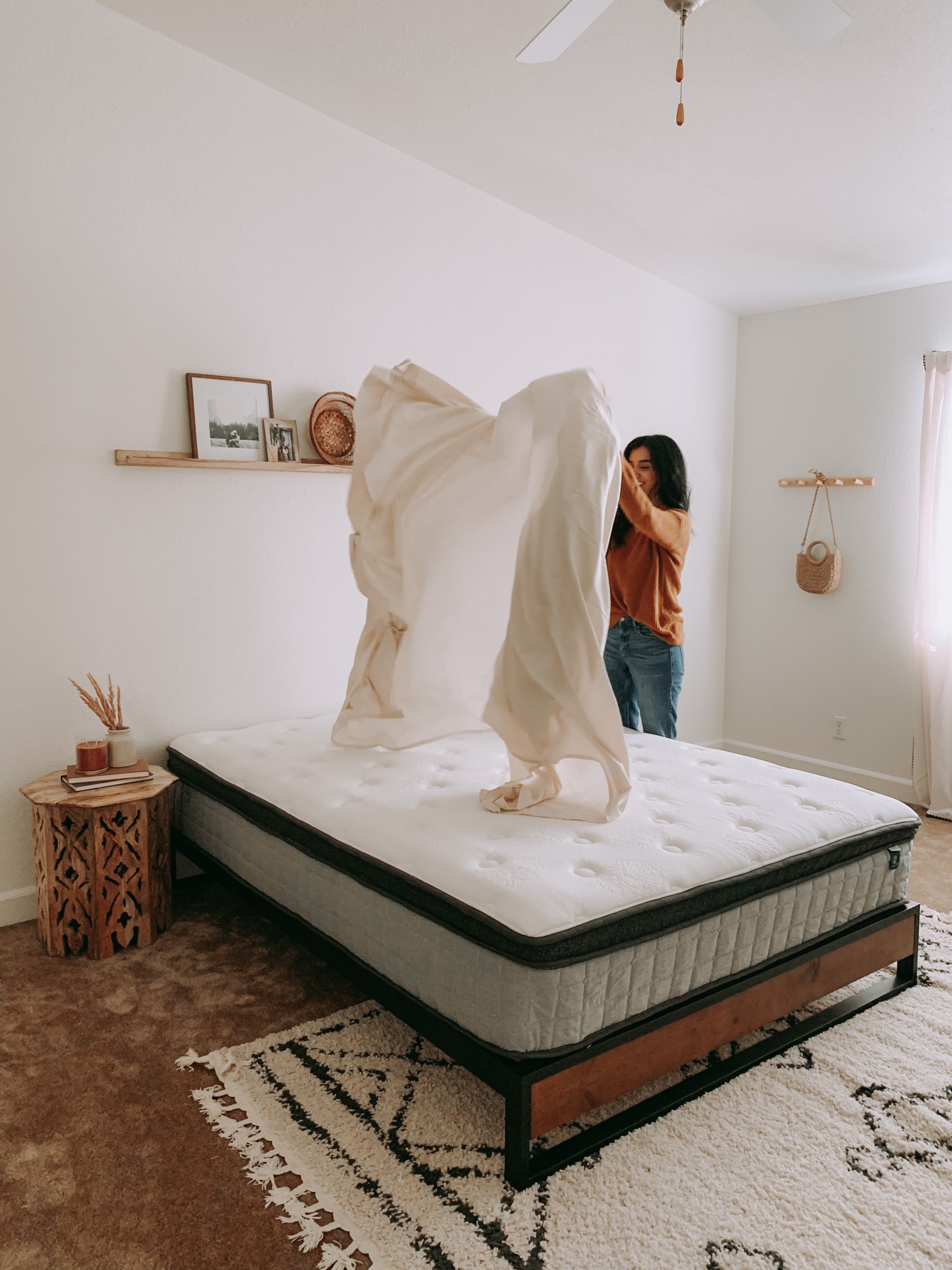Bedroom scene showing a person spreading a sheet over a Zinus hybrid mattress on a wood-and-metal frame, emphasizing easy setup and a relaxed, natural space with warm tones and minimal decor.