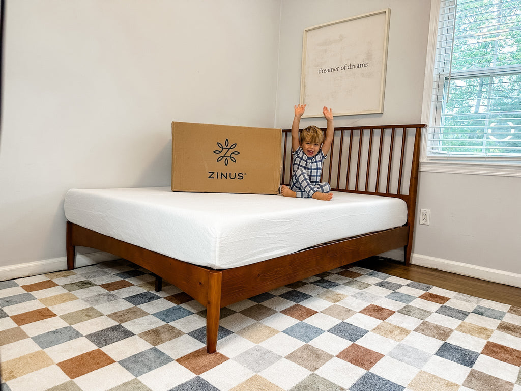 Child sitting on a new Zinus mattress beside a shipping box, raising arms excitedly.
