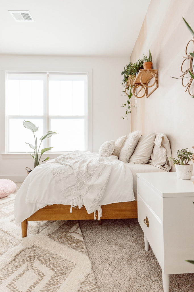 Bright bedroom with white bedding, wood bed frame, and plants on a softly patterned wall.