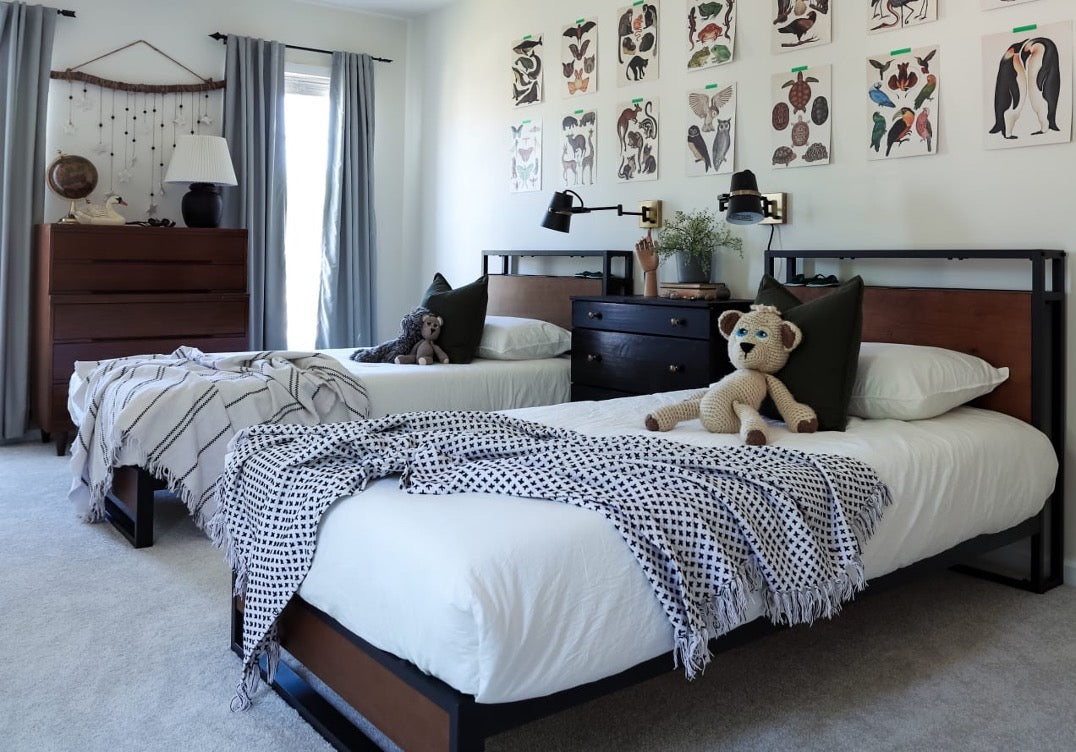 Kids’ bedroom with two matching wood-and-metal beds, white bedding, patterned throws, stuffed animals, and a gallery wall of playful animal prints