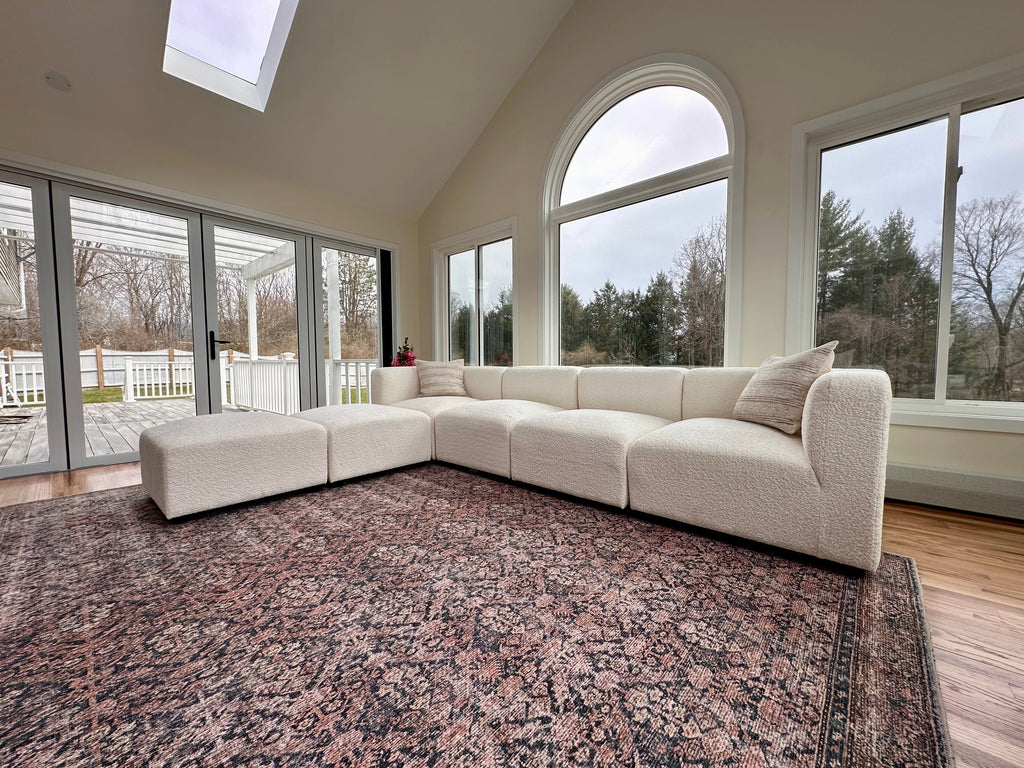 Spacious sunroom with a large cream sectional sofa, oversized windows, and a patterned rug on wooden floors