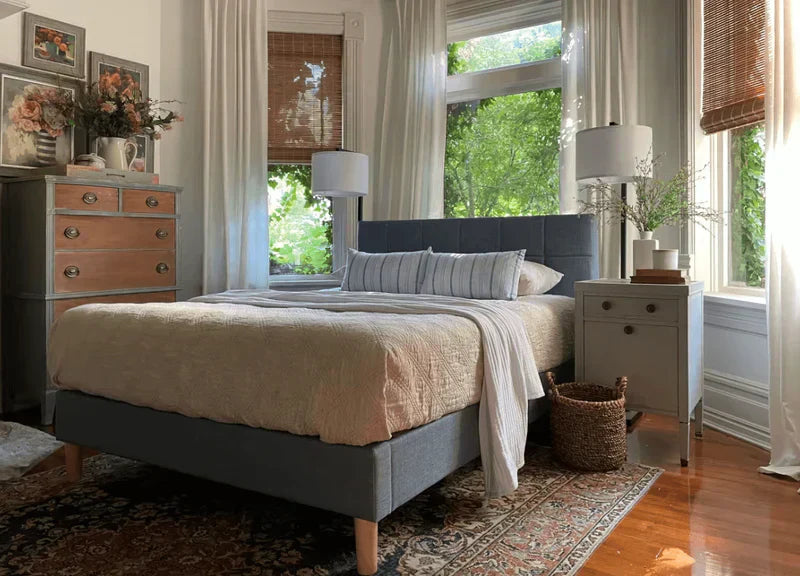 Sunlit bedroom featuring a blue upholstered bed, soft neutral bedding, layered pillows, tall lamps, and surrounding windows with sheer curtains and leafy outdoor views.