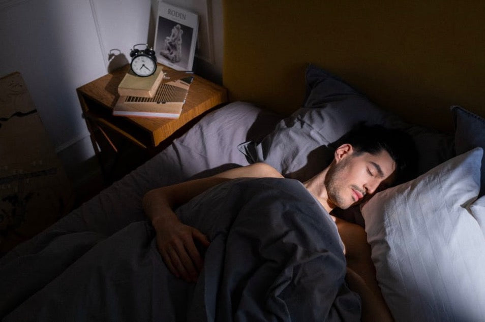 Person sleeping on a bed with soft gray bedding in a dimly lit room, shown beside a nightstand with books and an alarm clock to convey a calm, restful sleep setting