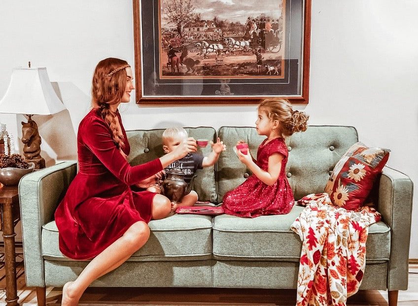 Person sitting on a light green sofa with two children, sharing playtime in a cozy living room styled with a framed painting, patterned pillows, and a warm, homey atmosphere