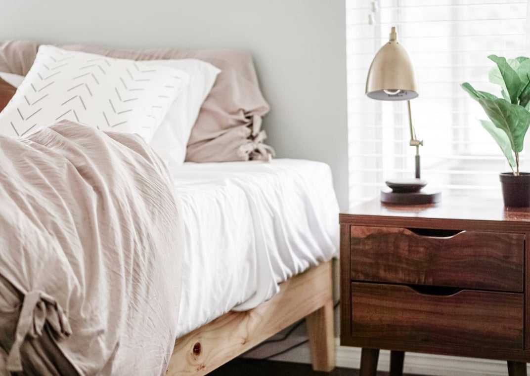 Minimal bedroom corner with a light wood bed, white bedding, soft neutral pillows, and a warm wood nightstand topped with a modern lamp and plant.