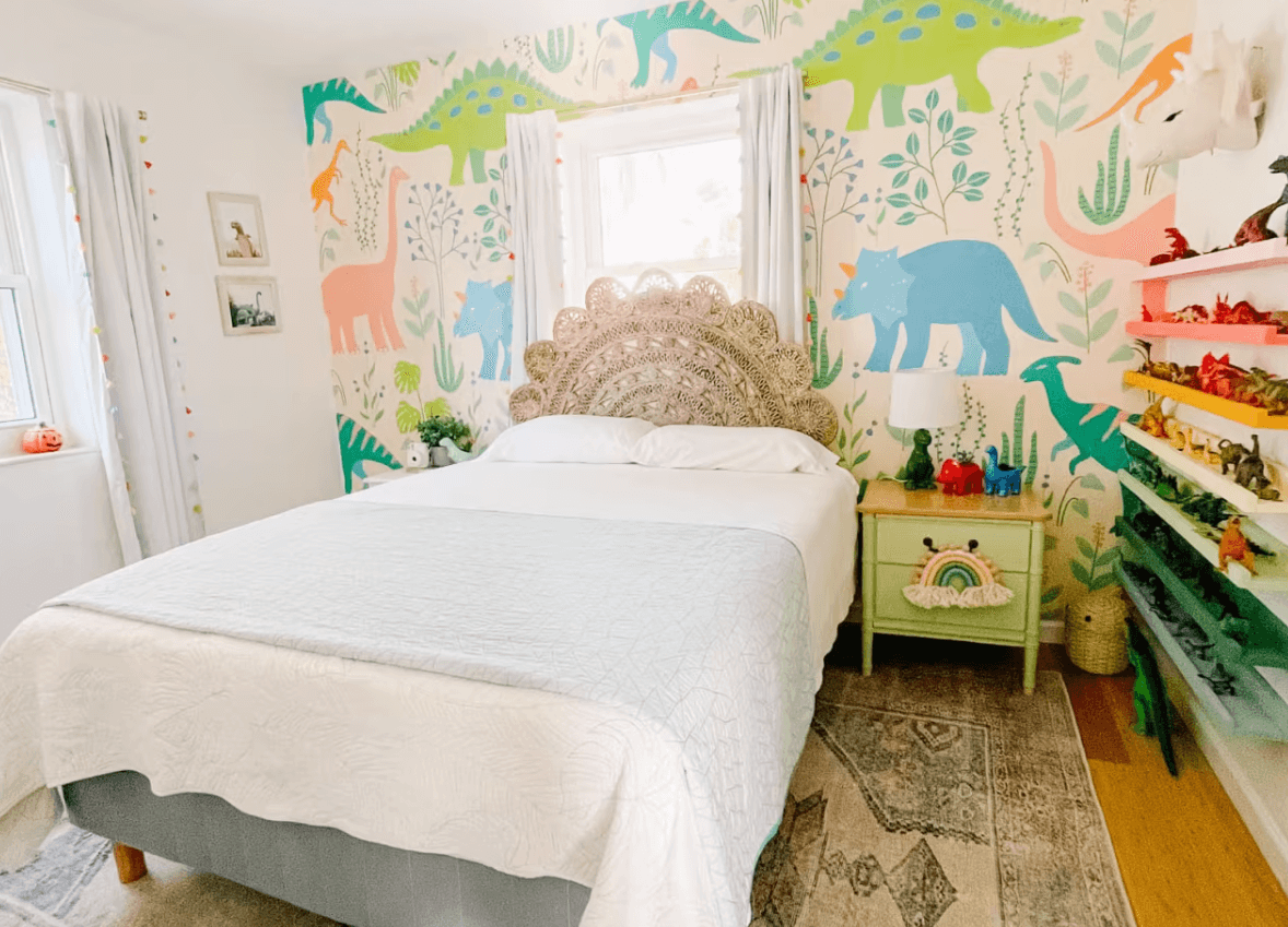 Colorful kids’ room with a carved headboard, white bedding, and a vibrant dinosaur mural, styled with playful decor and bright shelving.