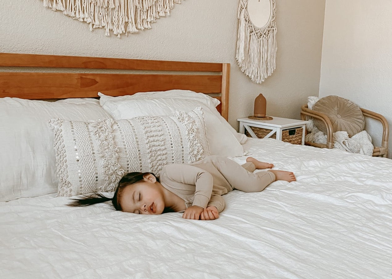 Small child sleeping on a white bed with textured pillows and soft neutral decor, creating a calm, cozy bedroom setting with natural woven accents