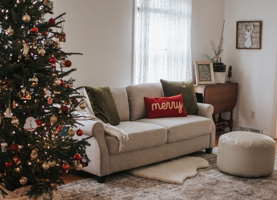 Living room decorated for Christmas with a lit tree, neutral sofa, green pillows, and a red “merry” pillow, creating a cozy holiday atmosphere.