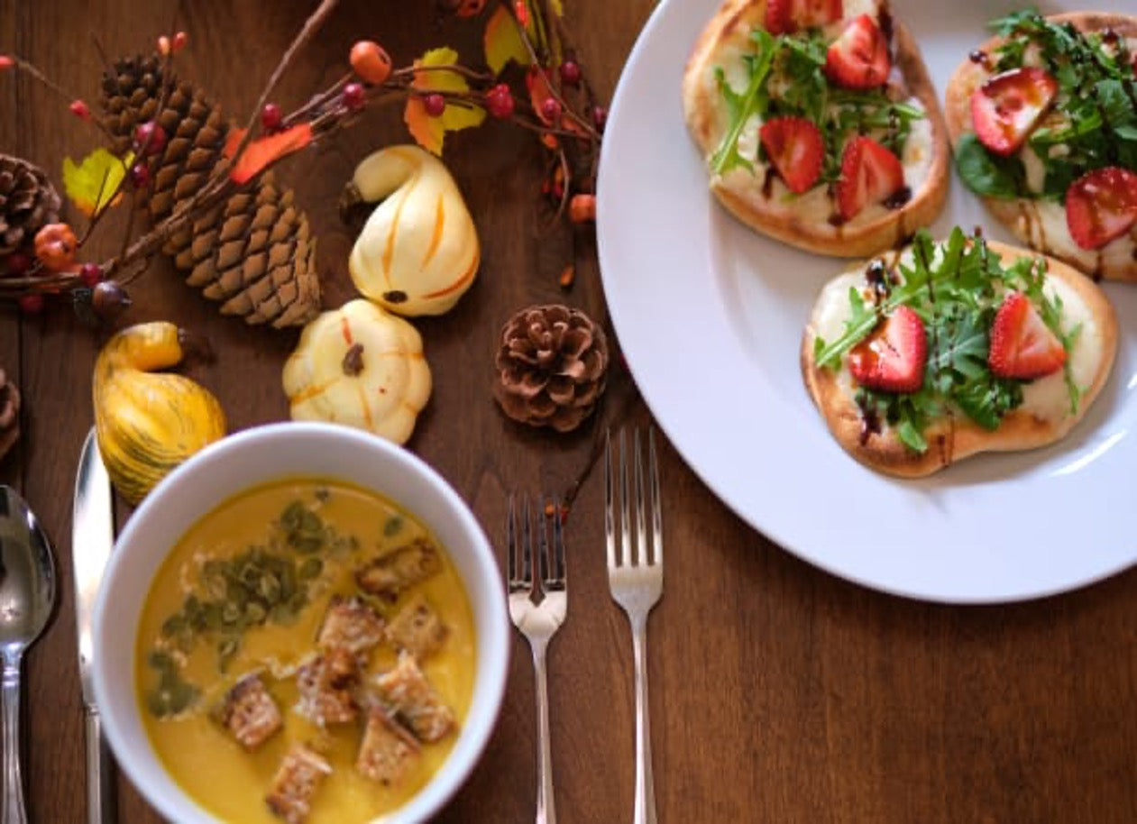 Fall-themed meal with a bowl of creamy soup topped with croutons and plates of flatbreads with strawberries and greens, set among pinecones and small gourds.