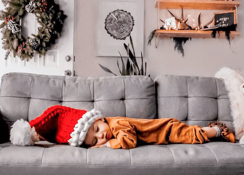 Child asleep on a gray tufted sofa wearing a festive red Santa hat, surrounded by holiday decor including a wreath and a decorated wall shelf.