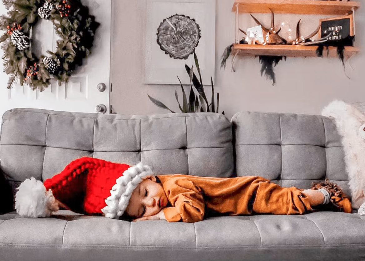 Child asleep on a gray tufted sofa wearing a festive red Santa hat, surrounded by holiday decor including a wreath and a decorated wall shelf.