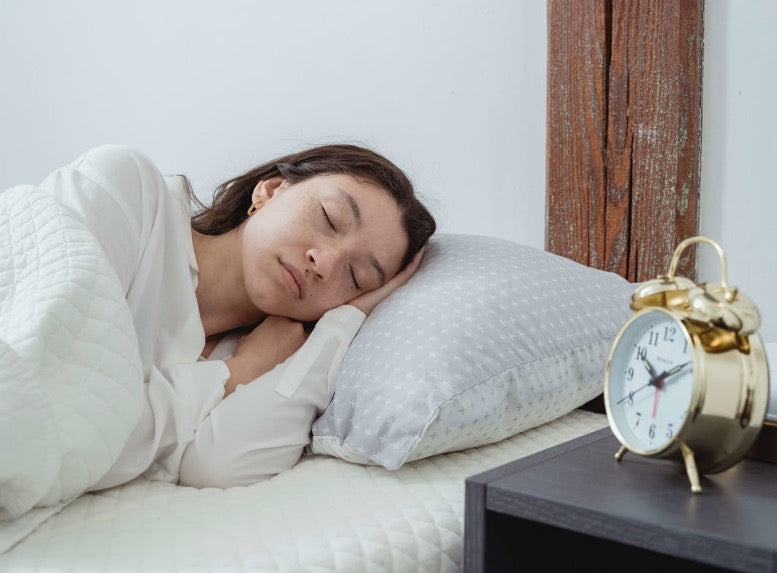 Person sleeping on a bed with white quilted bedding and a patterned pillow beside a bedside table with a gold alarm clock