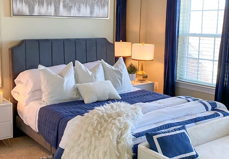 Bedroom styled with a gray upholstered bed, crisp white pillows, and navy bedding, accented by gold lamps and dark curtains for a polished, modern look.