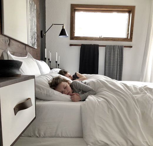 Two children sleeping peacefully on a bed with white bedding in a cozy room with wood accents, wall sconces, and layered blankets