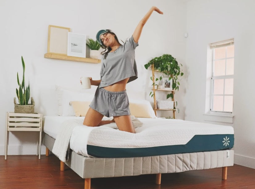 Person stretching on a mattress in a bright bedroom, holding a cup and wearing sleepwear, highlighting a refreshed wake-up moment with plants and soft decor
