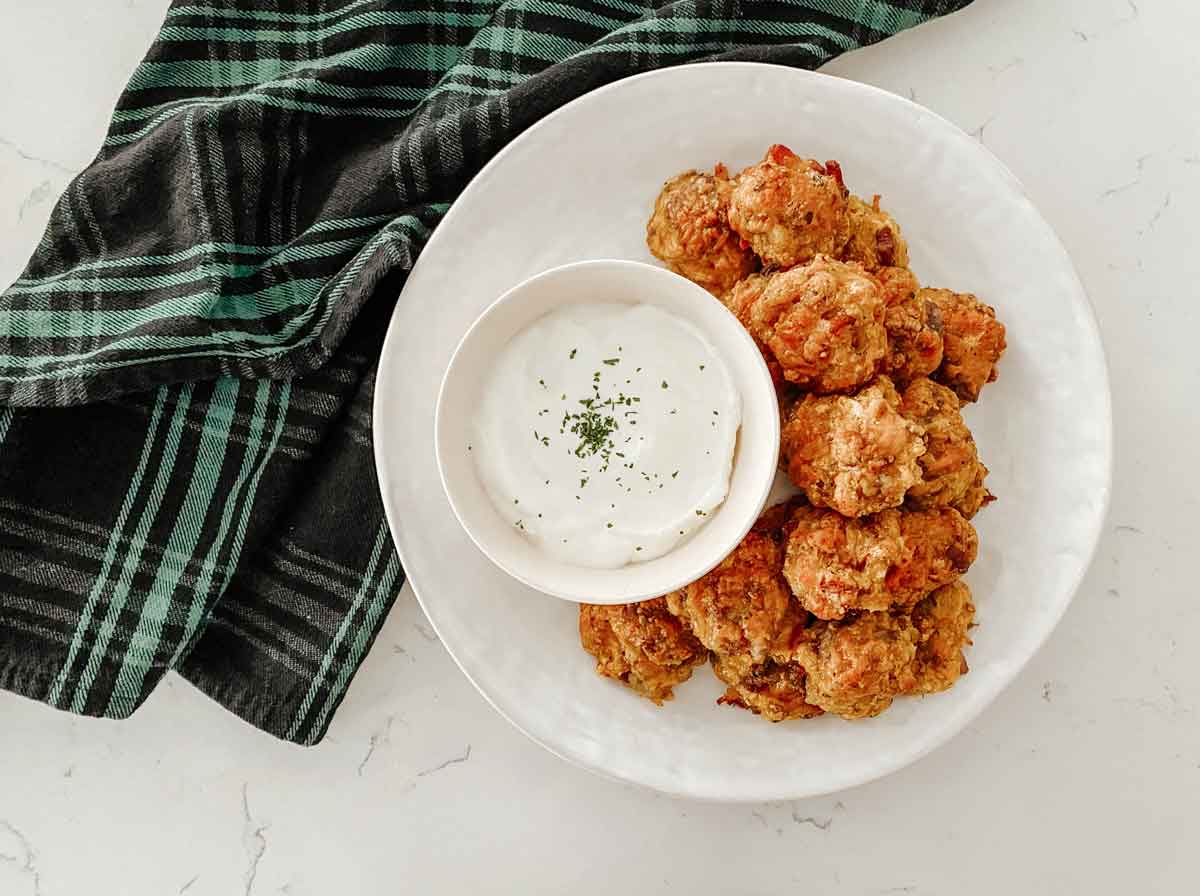 Plate of crispy fried bites served with a bowl of creamy dipping sauce, set on a white surface with a green plaid towel.