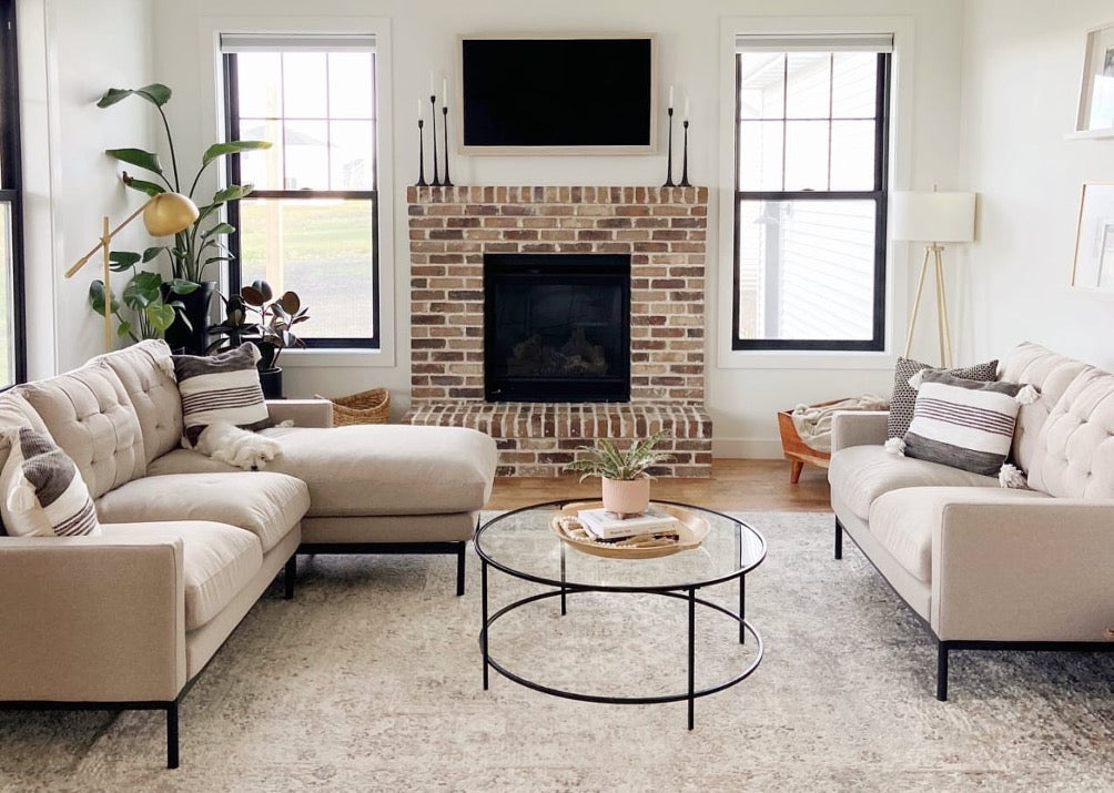 Bright living room with two beige tufted sofas, a brick fireplace, black-framed windows, and a round glass coffee table, styled with plants and neutral accents for a clean, modern feel.