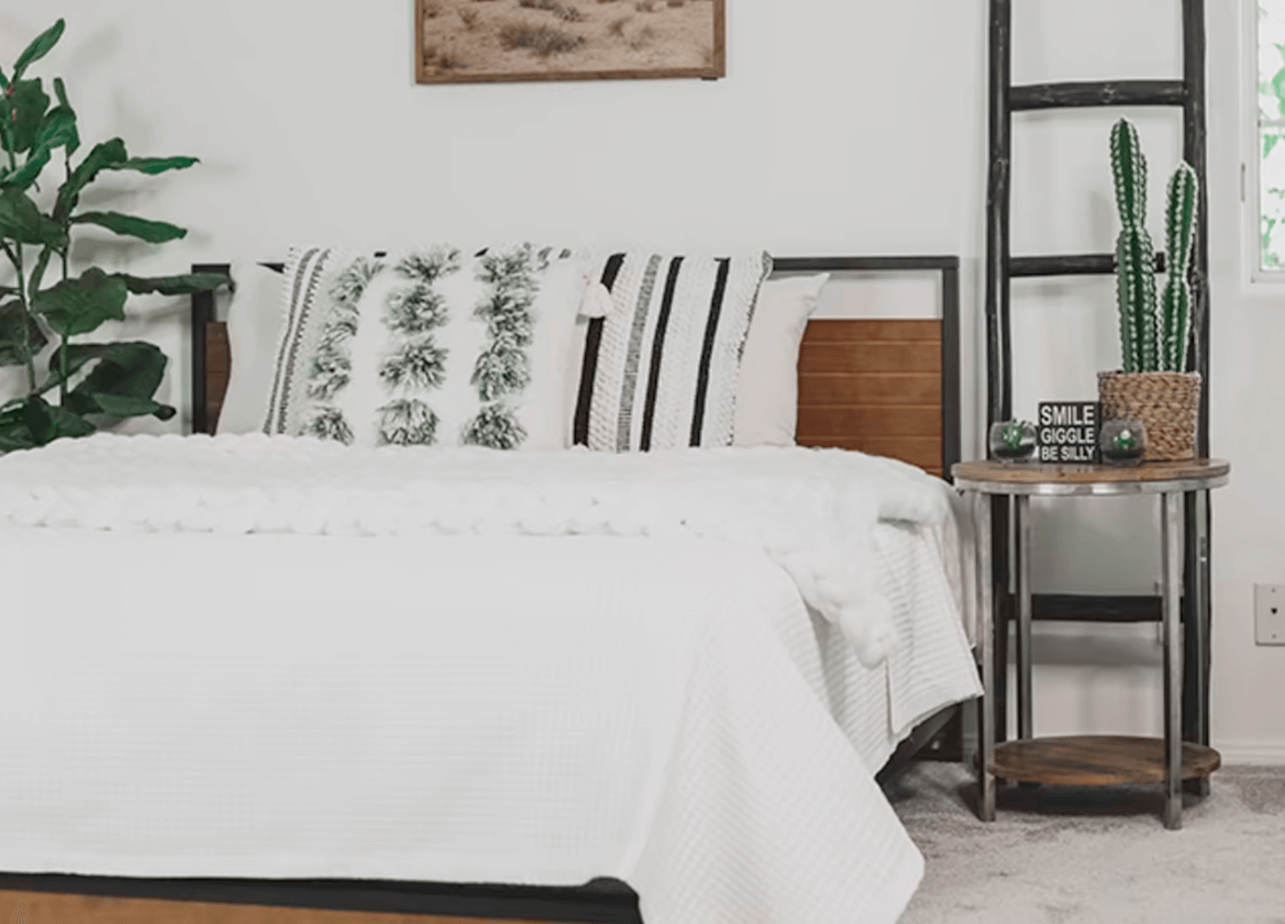 Cozy bedroom with a metal-and-wood bed frame, white bedding, textured black-and-white pillows, a small round side table, cactus décor, and a decorative ladder.