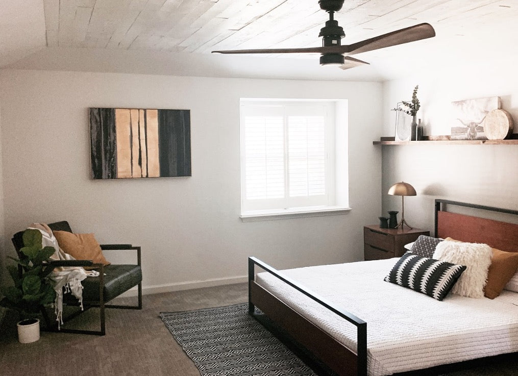 Neutral bedroom with a wood-and-metal bed frame, textured bedding, a dark rug, and a reading corner, styled with warm accents and a ceiling fan for a calm, modern look.
