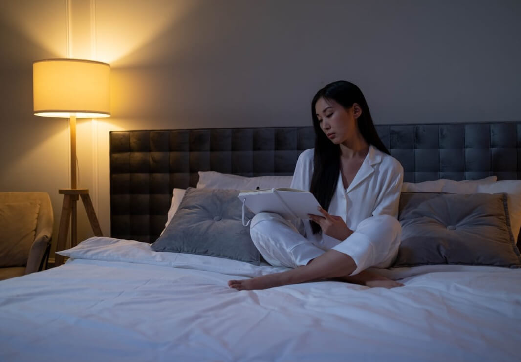 Person sitting on a bed at night reading from a tablet, surrounded by soft lighting, gray pillows, and a calm modern bedroom atmosphere