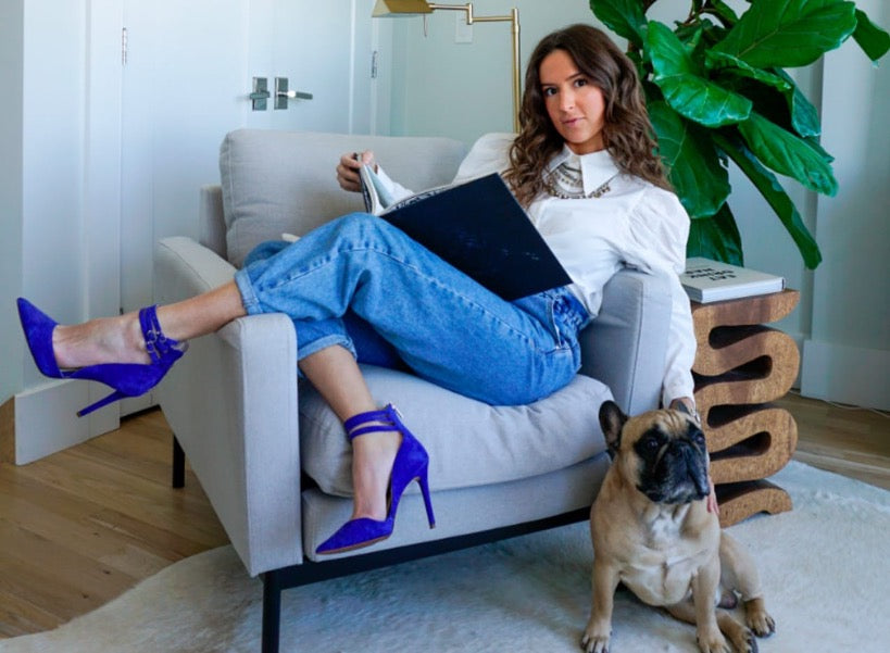 Person lounging on a modern armchair with a book while a small dog sits beside them, creating a relaxed, stylish corner with bold heels and a large plant.