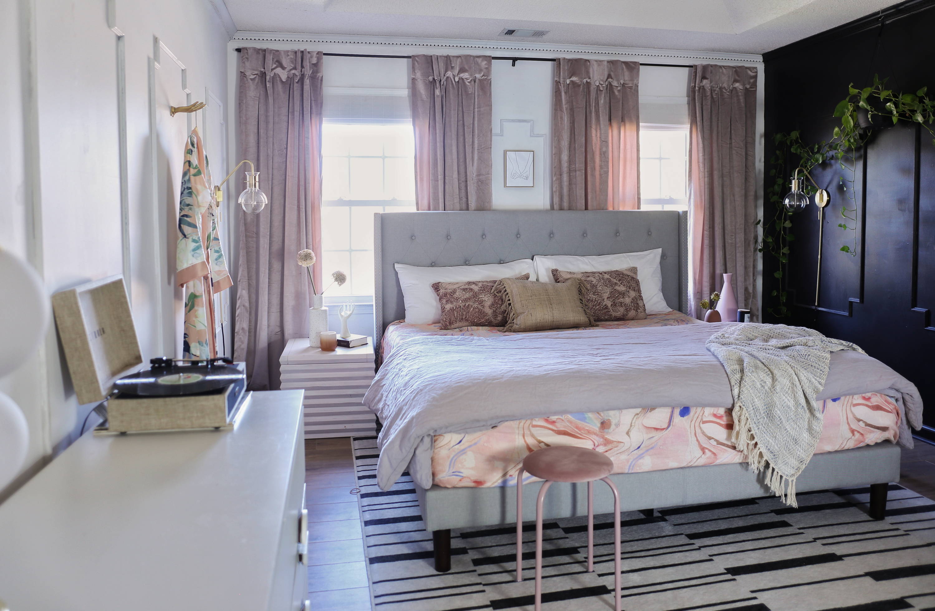 Stylish bedroom with a gray upholstered bed, layered bedding, soft pink accents, striped rug, and tall curtains framing large windows.
