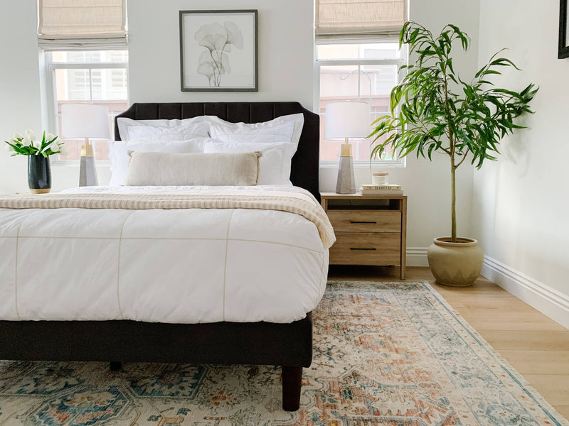 Bright bedroom with a dark upholstered bed, layered white bedding, soft neutral pillows, wood nightstands, and a tall potted plant for a fresh, airy look