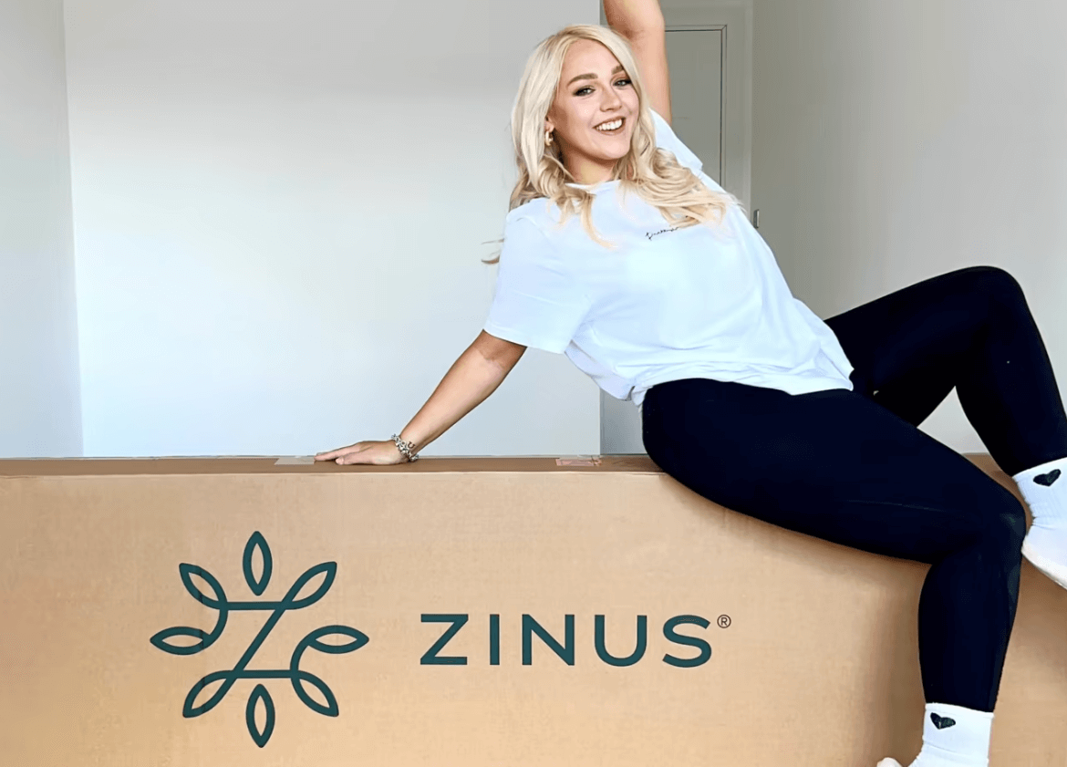 Smiling woman sitting playfully on a large Zinus mattress box in a bright, empty room.