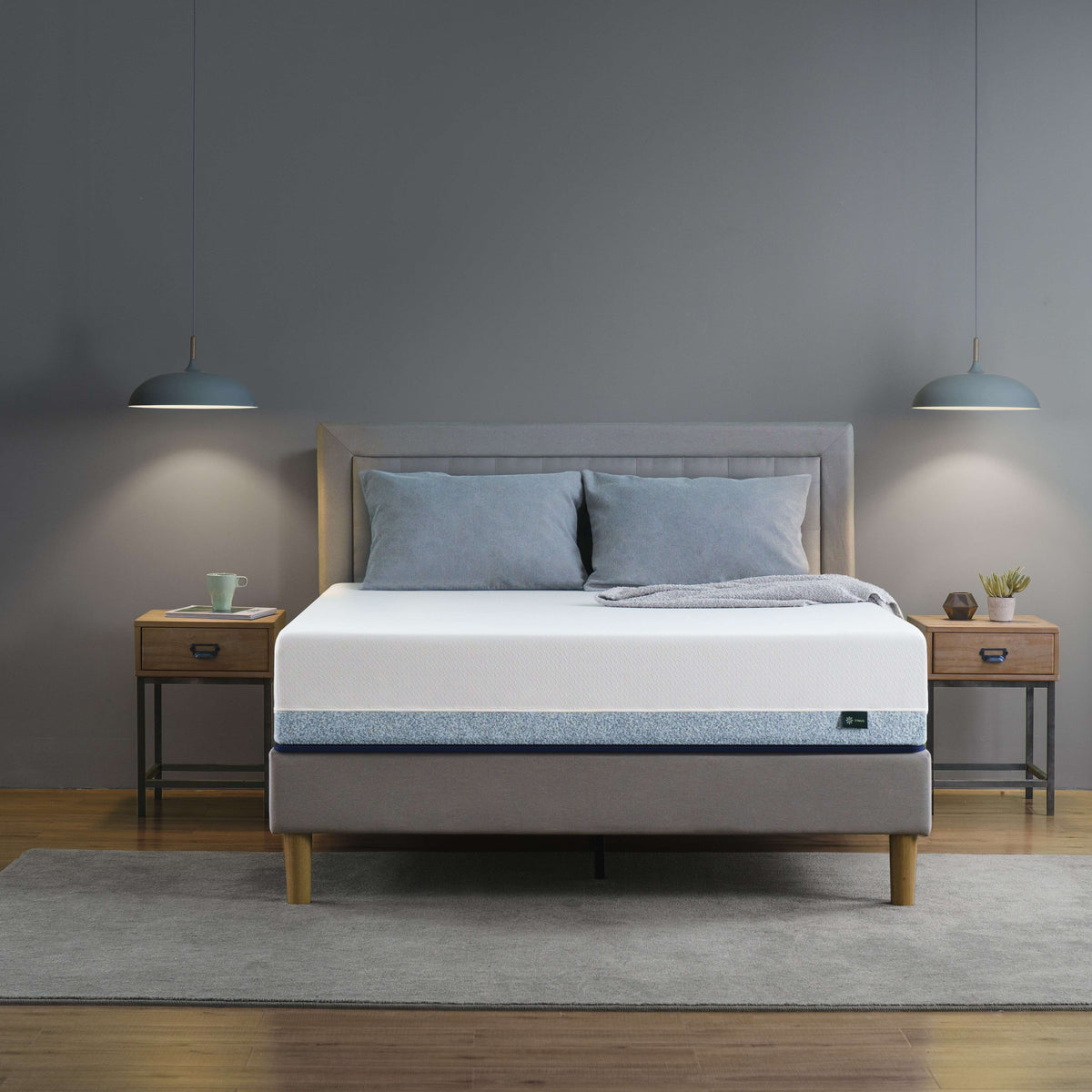 Zinus Original Green Tea Hybrid Mattress styled in a modern bedroom with gray upholstered headboard, wooden legs, and two nightstands. Mattress features a white top with blue accent side panels, offering supportive comfort in a queen size design, ideal as a hybrid cooling mattress option.