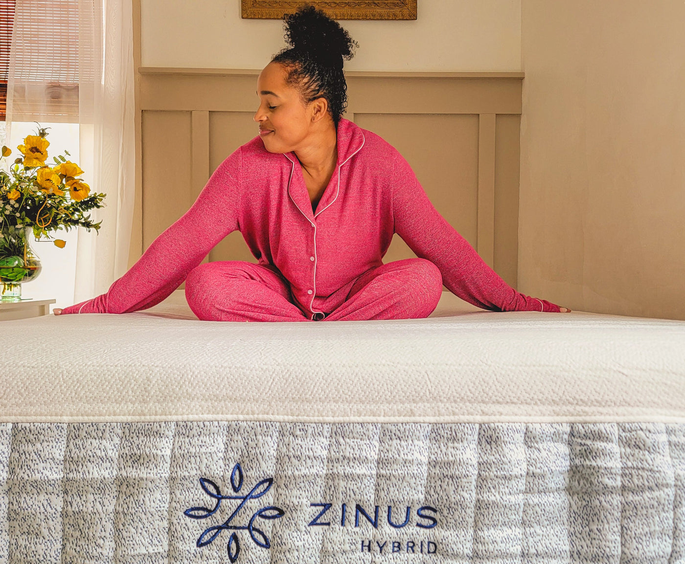 Person in bright pink pajamas sits cross-legged on a Zinus Hybrid mattress with white quilted top and gray patterned side panel featuring Zinus logo, styled in a cozy bedroom with light walls, framed art, and fresh yellow flowers on the nightstand