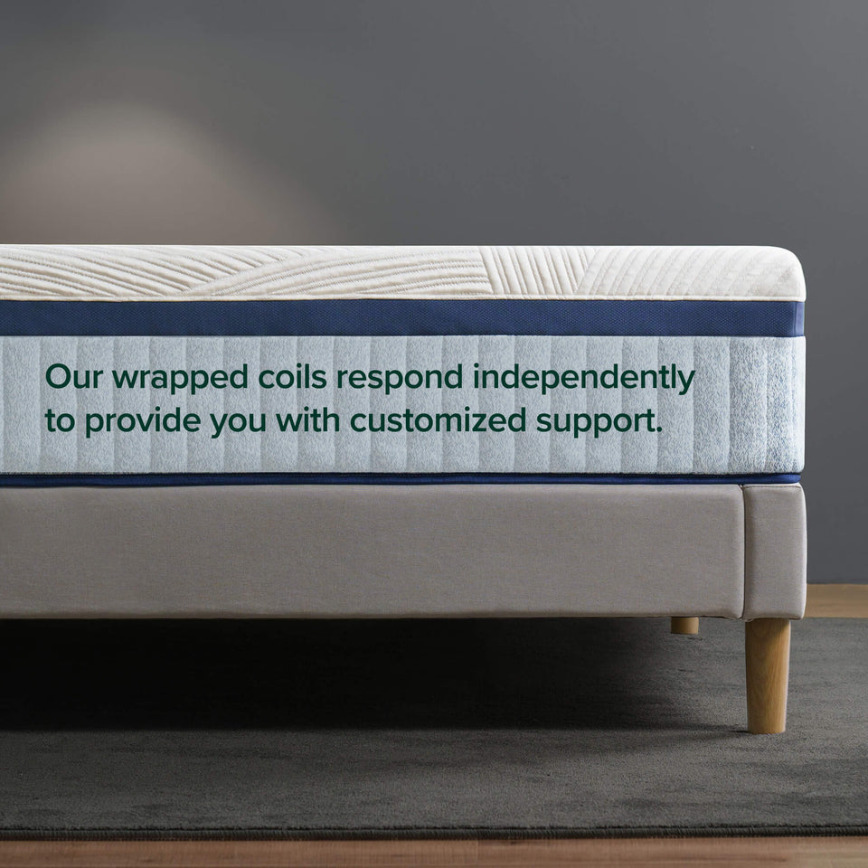 Zinus Ultra Cooling Green Tea Hybrid mattress edge close-up showing wrapped coil and gray foam layers beneath white cooling cover, with text: Our wrapped coils respond independently to provide you with customized support, on gray upholstered bed frame with wood legs.
