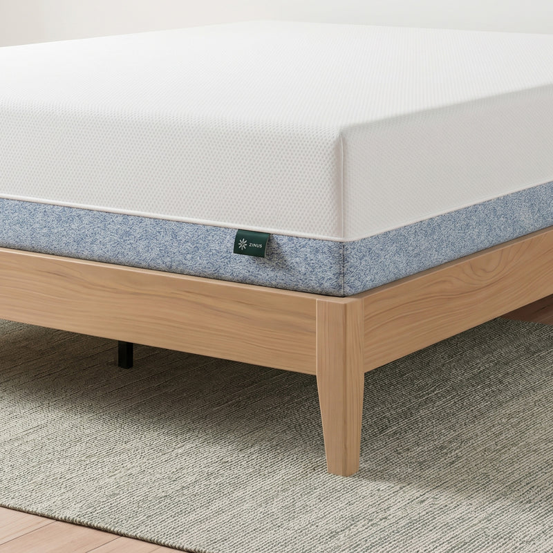 Zinus Balanced Comfort Core Memory Foam Mattress, close-up corner view showing the white knit top cover, blue woven side panel, and Zinus brand tag, set on a natural wood platform bed frame with visible under-bed clearance and clean tailored stitching along the seam