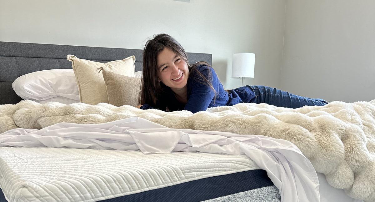 Smiling woman lying across a Zinus mattress with white top and gray side panel, styled with fluffy cream blanket, gray headboard, and neutral pillows in a bright bedroom, conveying comfort and relaxation in a cozy modern setting