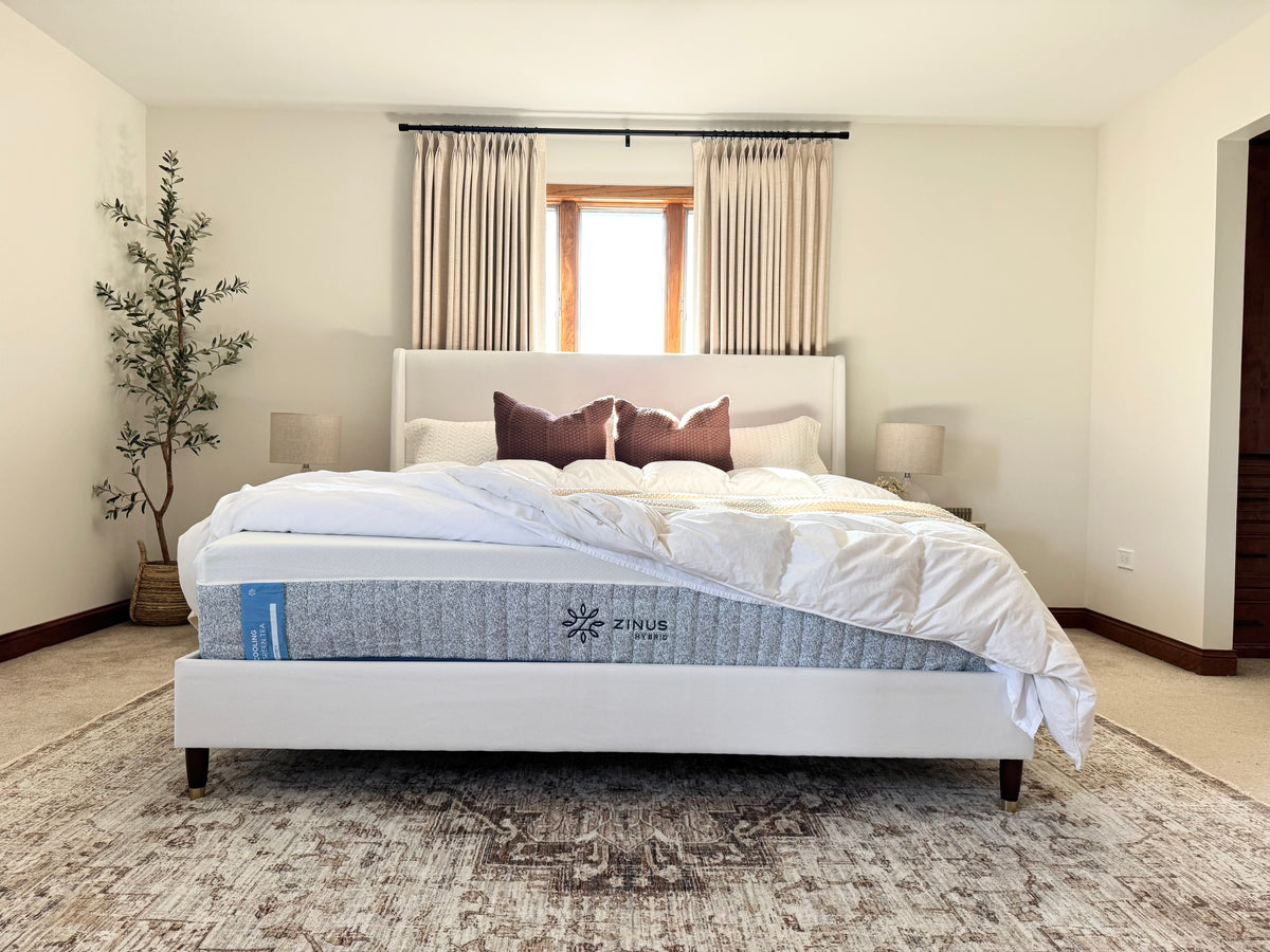 Stylish bedroom featuring a Zinus Hybrid mattress on a white upholstered frame with cream bedding and brown accent pillows. Sunlight filters through curtains, highlighting comfort, elegance, and inviting design for a modern restful space.