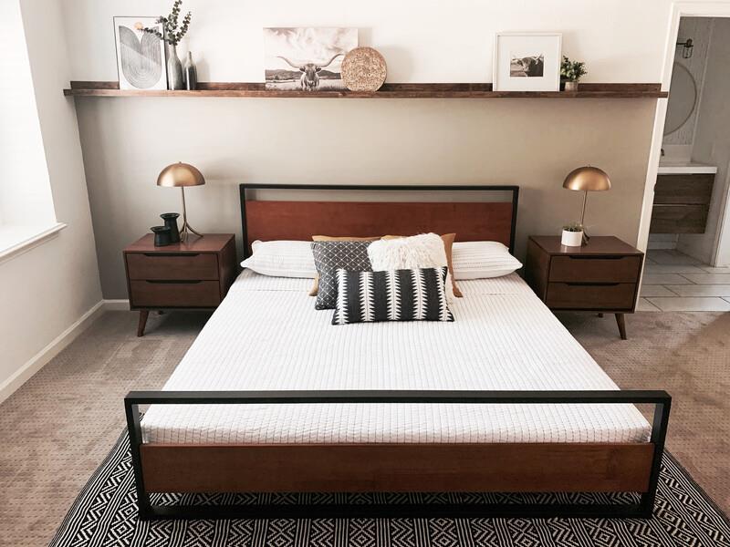 Mid-century modern platform bed with a wood headboard and black metal frame, dressed in white bedding and patterned accent pillows; styled with matching wood nightstands and brass dome lamps beneath a floating shelf to highlight sturdy support, minimalist design, and warm contemporary bedroom appeal.