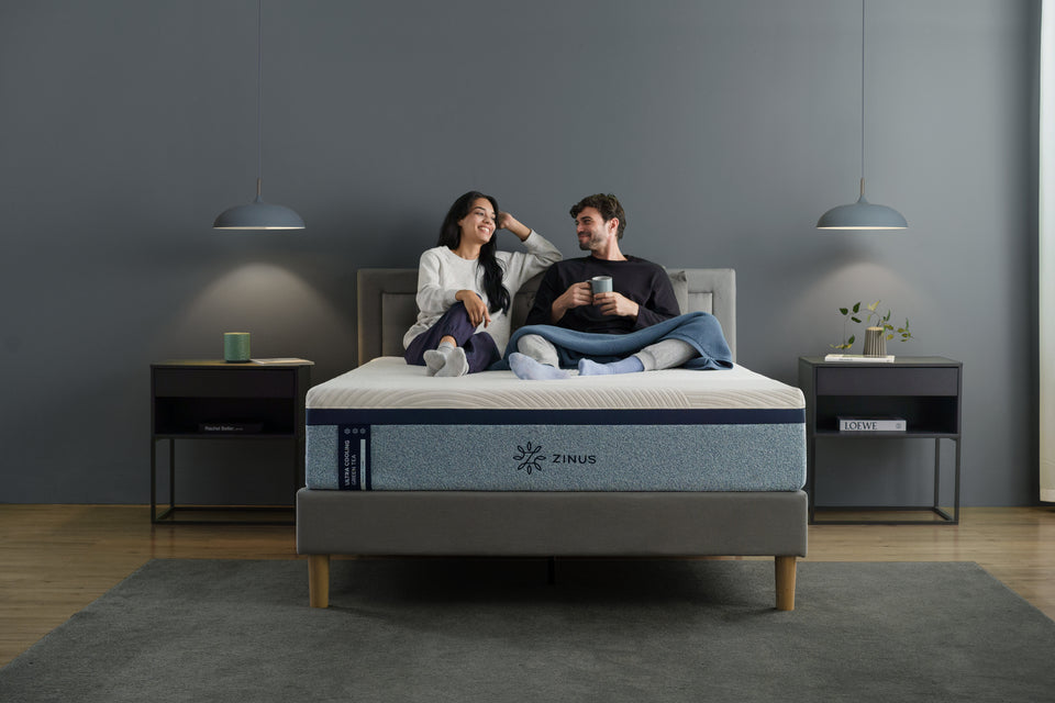 Happy couple sits together on a Zinus Ultra Cooling Green Tea mattress, wrapped in a blue blanket. Modern gray bedroom with nightstands and lamps. Lifestyle photo highlights mattress comfort, couple bonding, and premium sleep experience